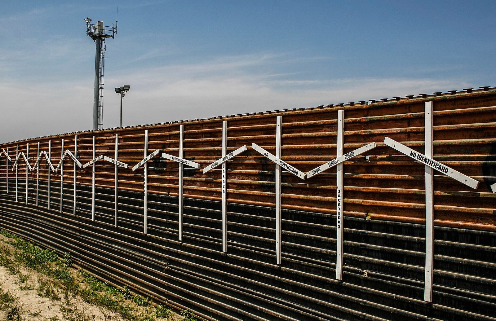 Memorial crosses on the border wall, marked with names and the words for the unidentified. The same boundary that carries the region's economy also carries a human cost.