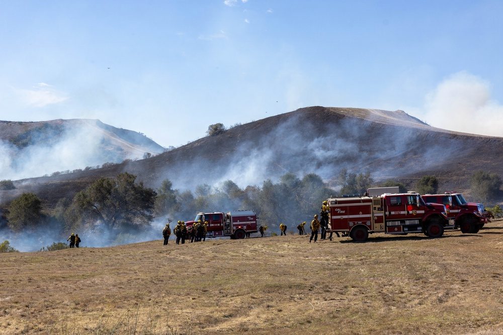 Cal Fire engines stage on a grass field at Camp Pendleton on Friday, October 16, as smoke rises from the blackened hills behind them. Crews first reached 20 percent containment on Friday evening.