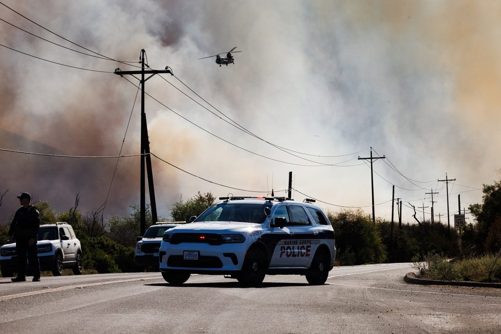 A Marine Corps Police vehicle blocks a road at Camp Pendleton on Thursday as smoke rises behind it and a helicopter crosses above. Authorities closed base roads including Las Pulgas Road while crews worked the fire.