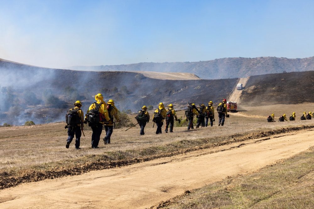 Firefighters walk a burned slope at Camp Pendleton on Friday, October 16, as light smoke rises behind them and an engine waits on the road above. Crews stayed on the fire through Friday as it crossed containment lines on its northwest side.