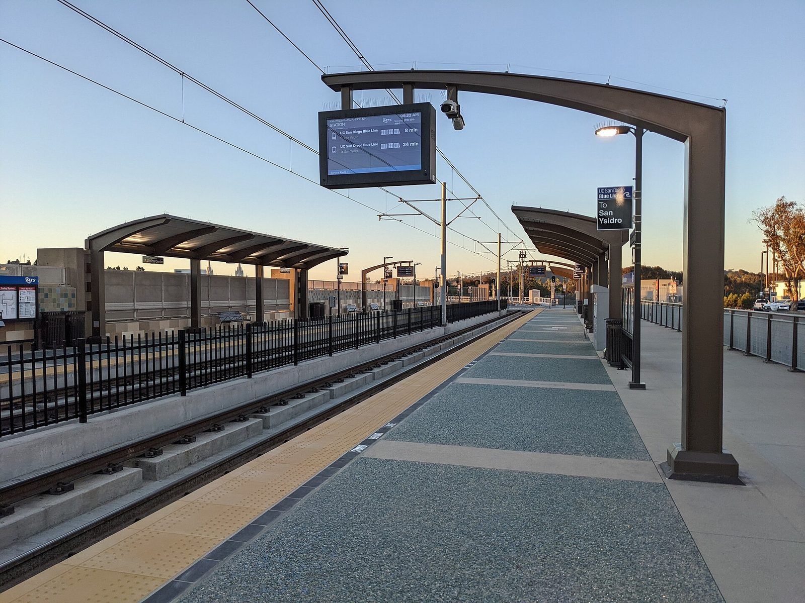 The VA Medical Center station platform at first light, the departure board showing Blue Line trains toward San Ysidro. For many patients the trip to an appointment starts here, which is why the state of the buildings at the top of the hill matters.